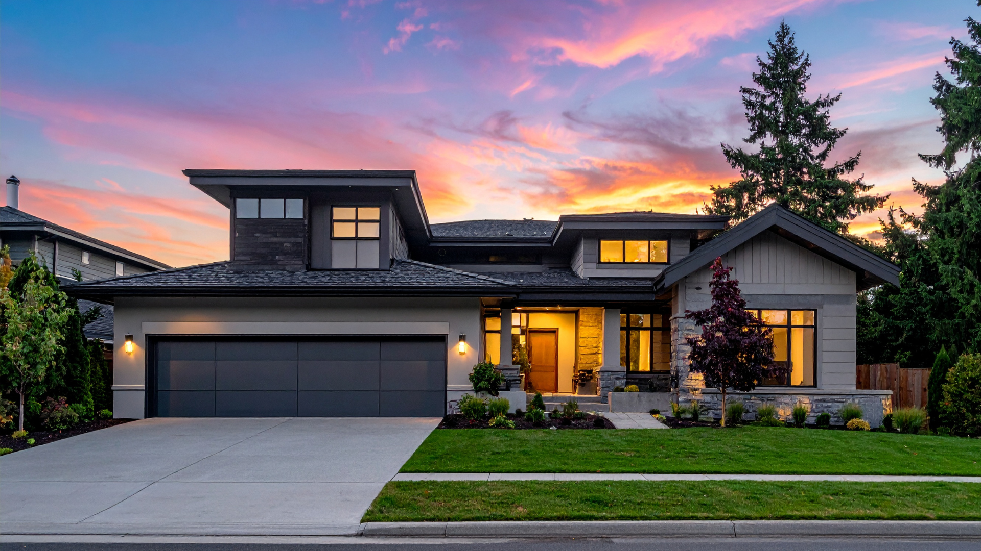 Modern home with sleek architectural design featuring a dark roof, large windows, and a stone-accented exterior, set against a colorful sunset sky.