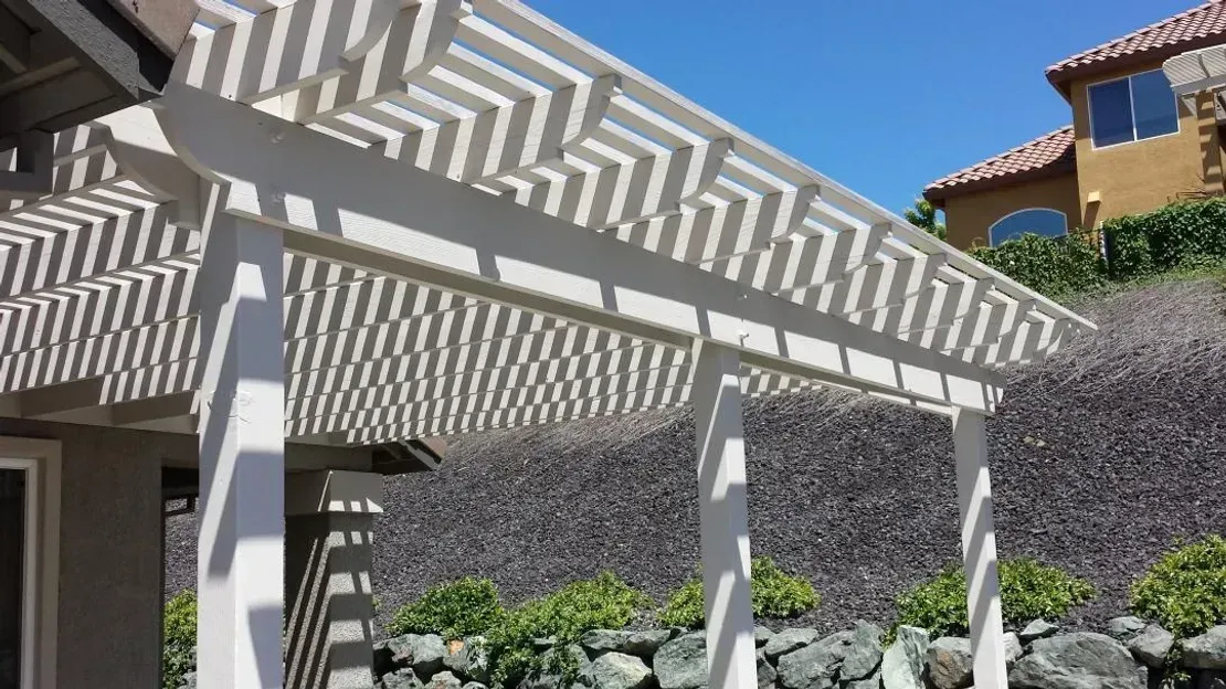 White pergola casting zig-zag shadows on a patio with a sunny blue sky.