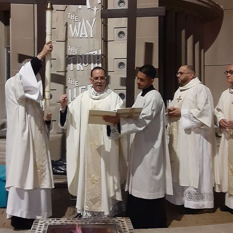 A group of clergy and altar servers dressed in white liturgical garments. One person holds a large lit candle while another holds an open book.