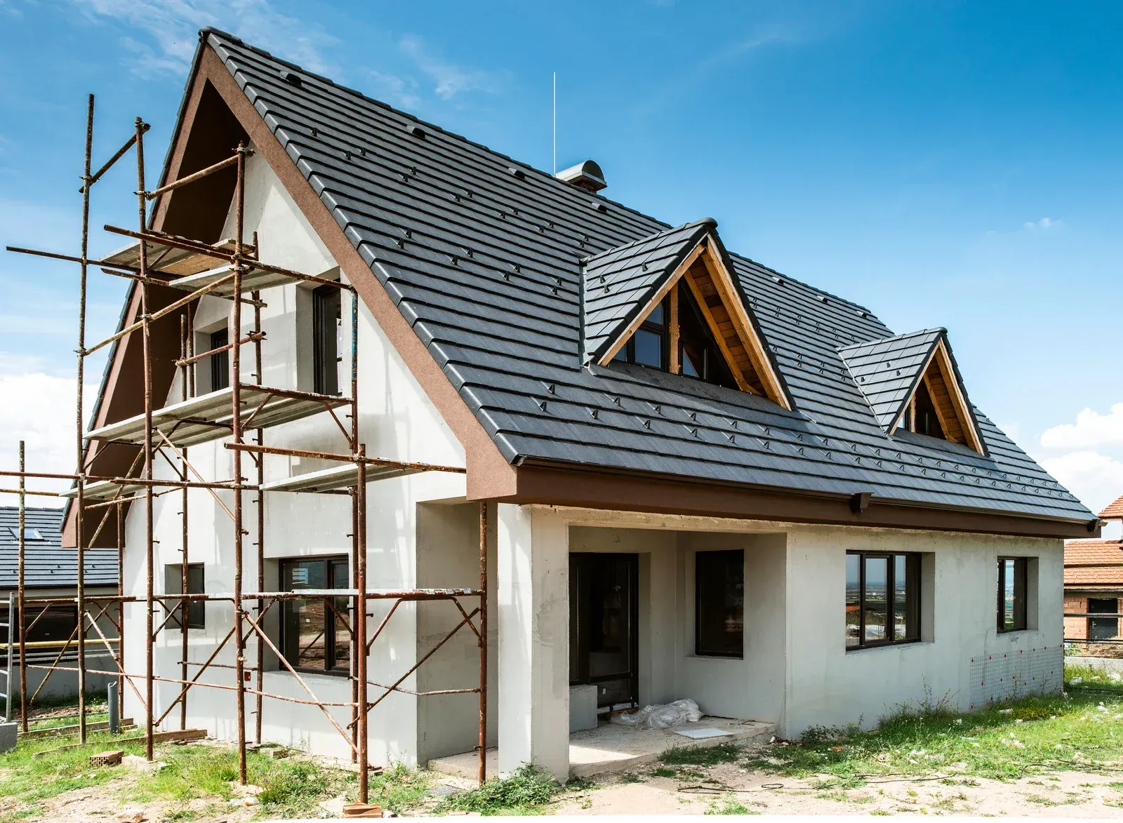 House under construction, gray stucco exterior, dark roof, scaffolding, blue sky.