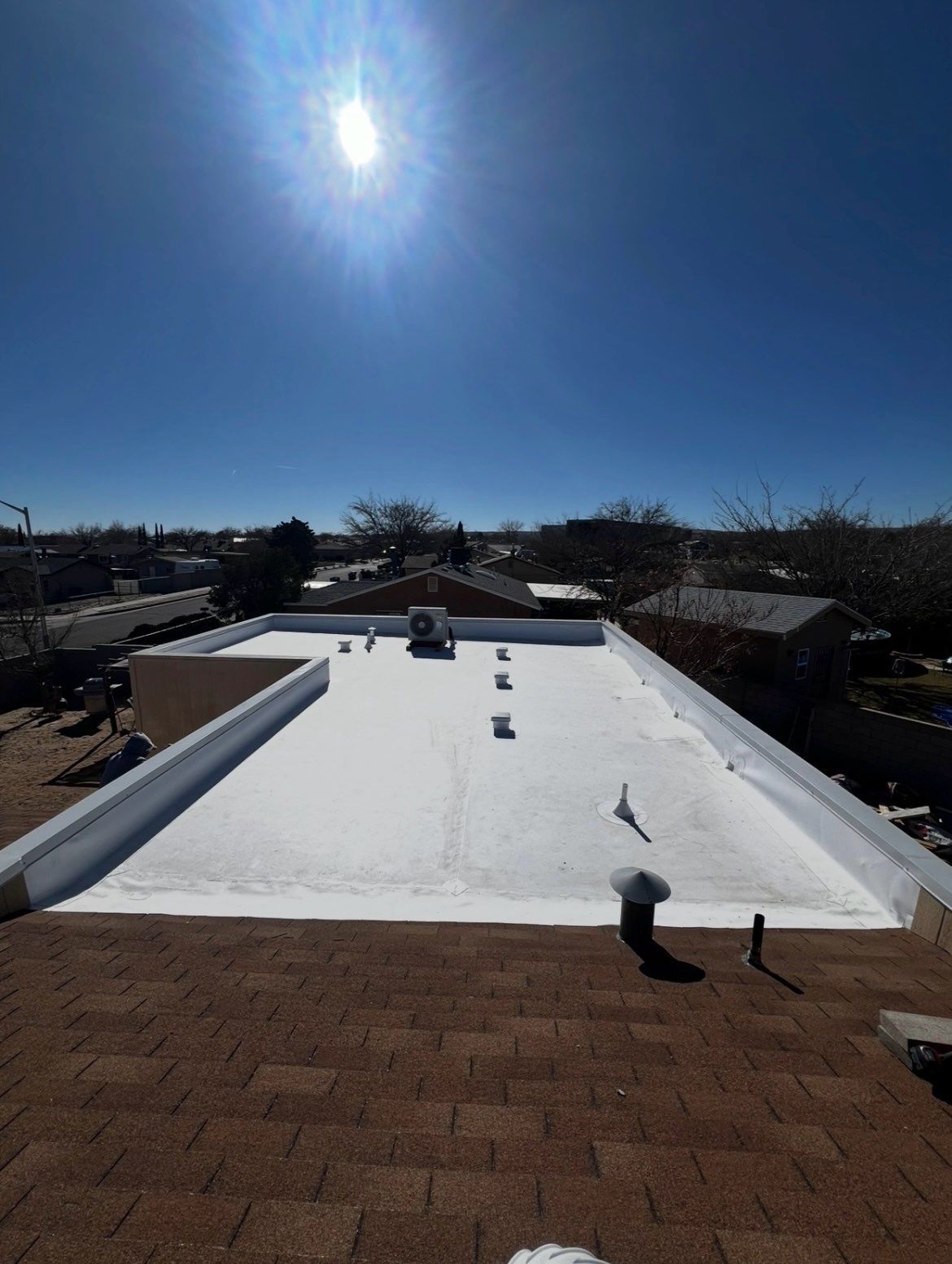 Beige house with brown roof and a blue sky. A patio with chairs is visible.