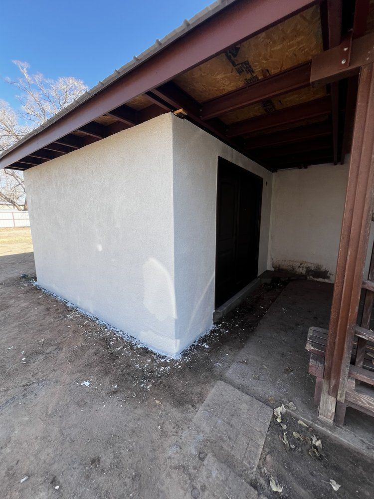 White stucco building with a dark door, beneath a brown awning, and a concrete ramp.
