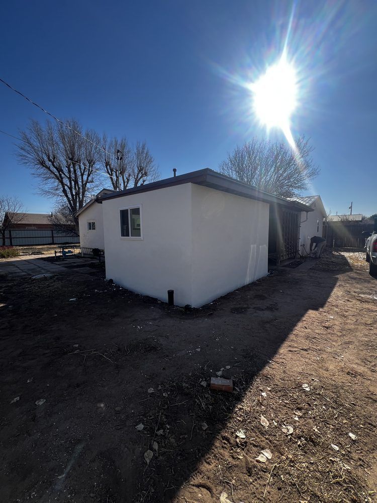 White stucco building with a window and a dark roof under a bright sun.