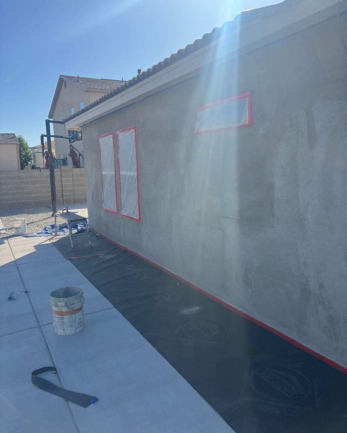 Exterior wall construction with windows taped off. Grey stucco, blue sky, and a sunny setting.