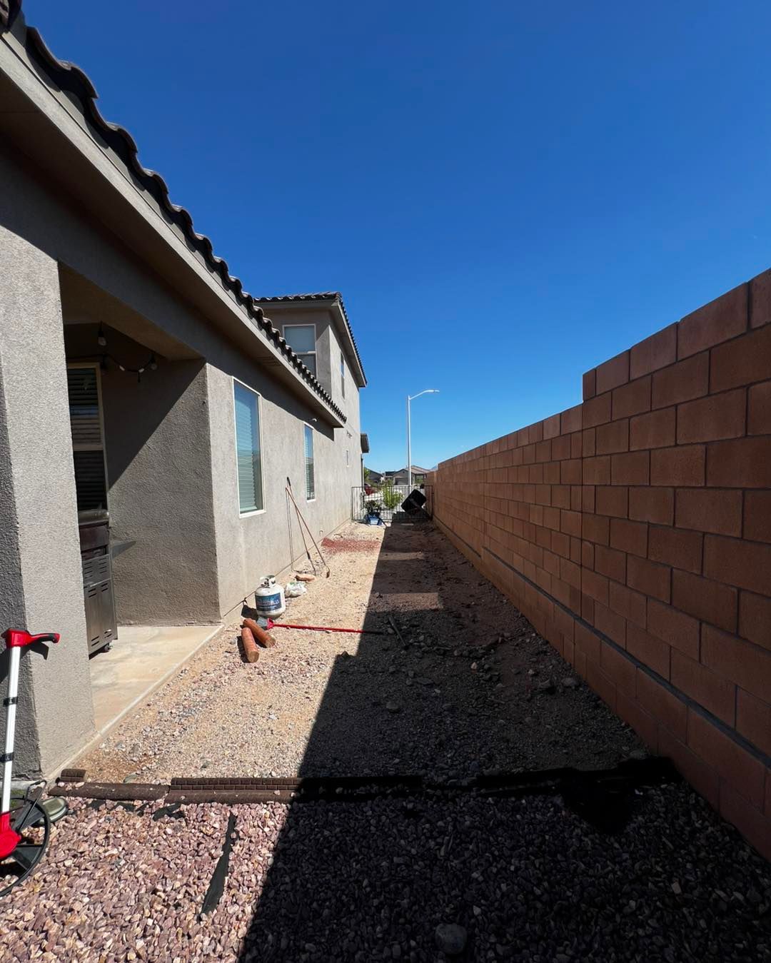 Narrow side yard with gravel, brick wall, and house. Blue sky overhead.