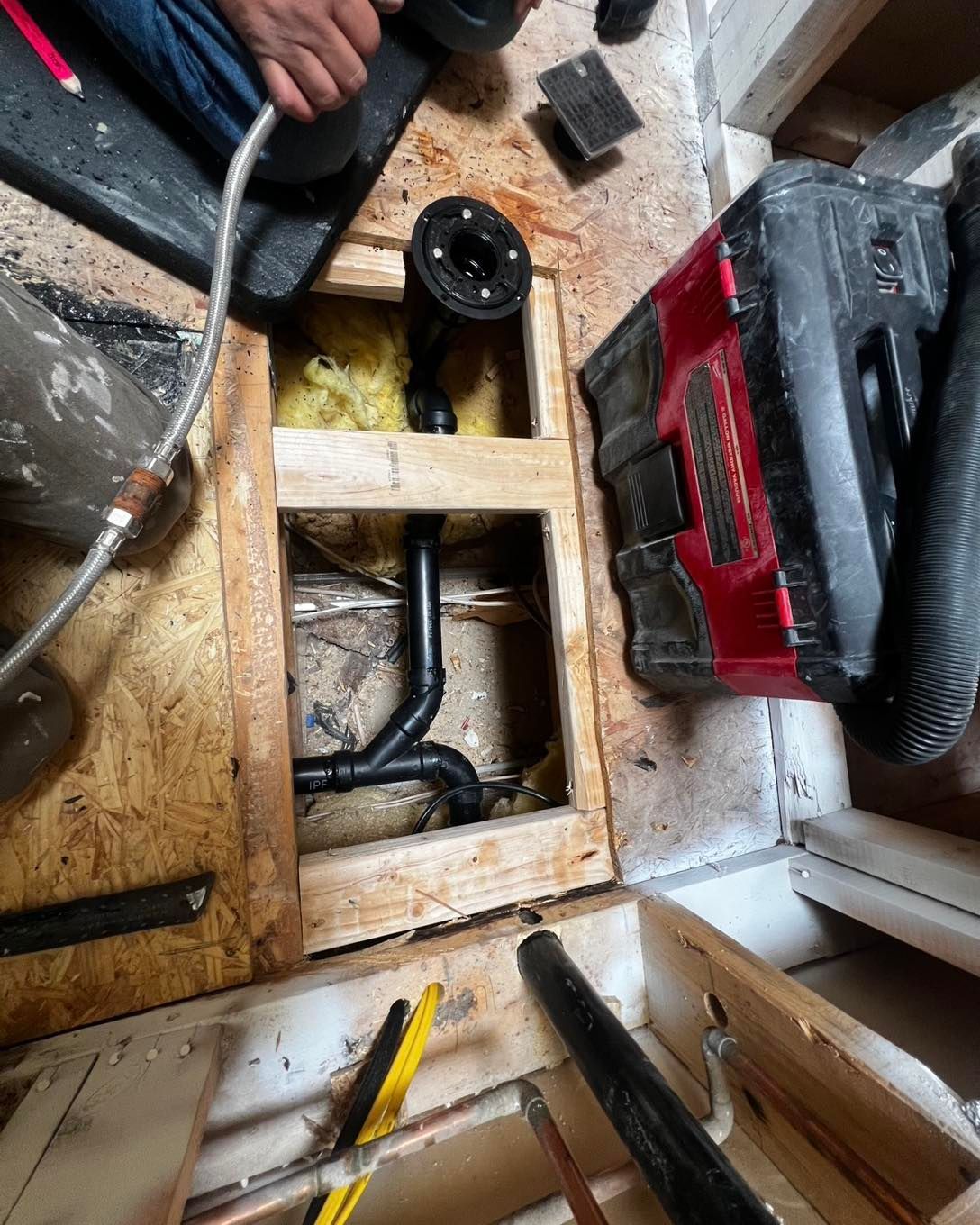Plumbing work in progress: Black pipes, shower drain, tools, and a red vacuum on a wooden floor.