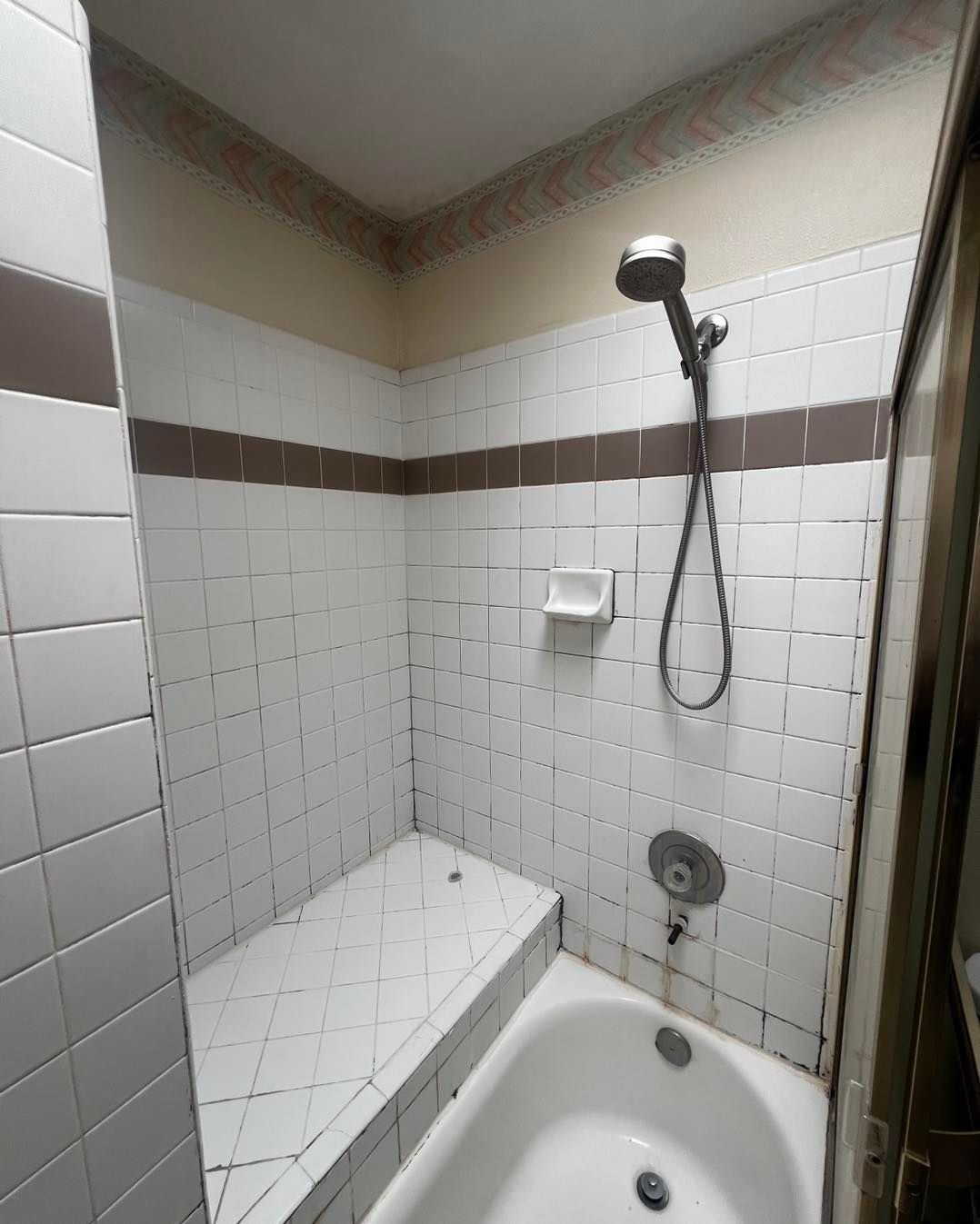 Shower stall with white tile, brown accent, built-in seat, and handheld showerhead.