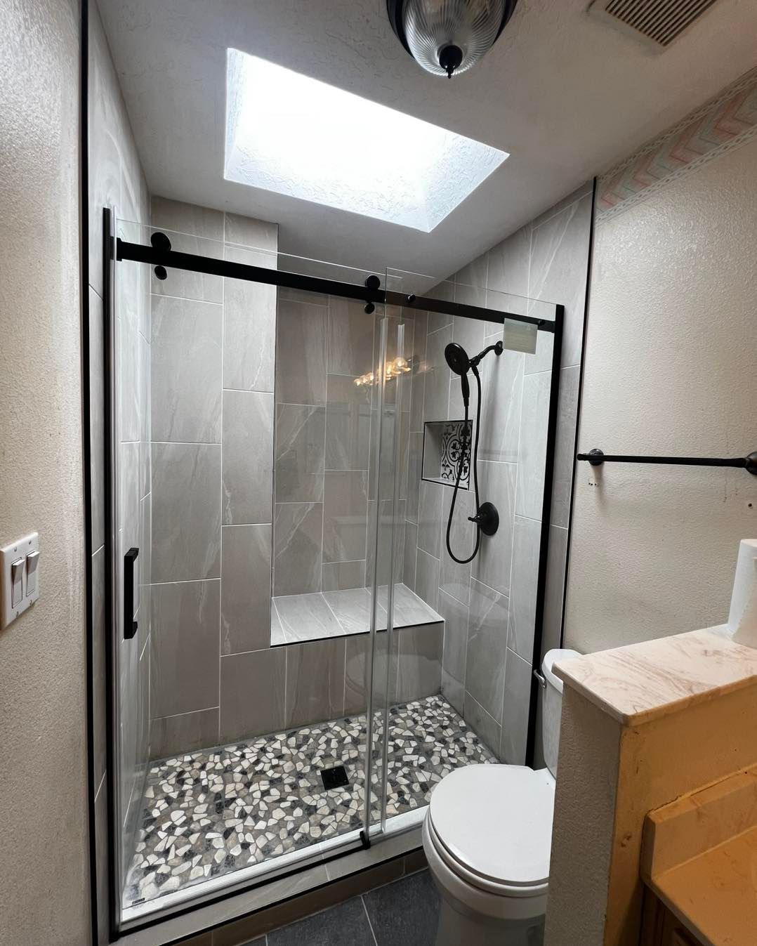 Modern bathroom with gray tiled shower, pebble floor, and sliding glass door. A skylight provides light.