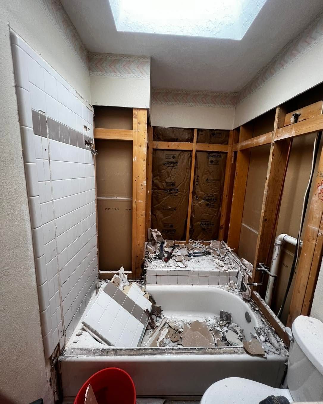 Bathroom undergoing renovation; exposed studs, demolished tile and tub, open walls, overhead skylight.