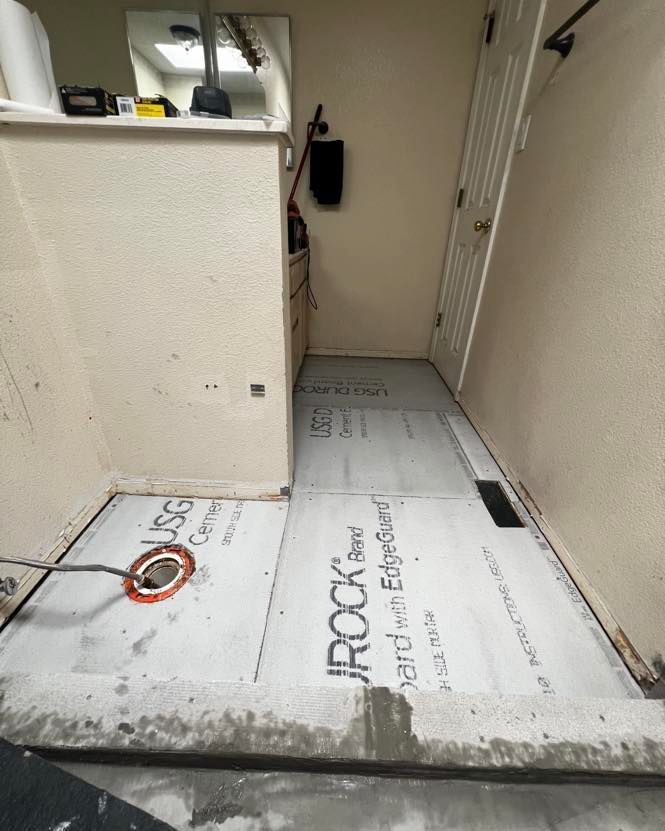 Shower floor renovation in progress: gray waterproofing boards laid out, drain visible. White walls and door.