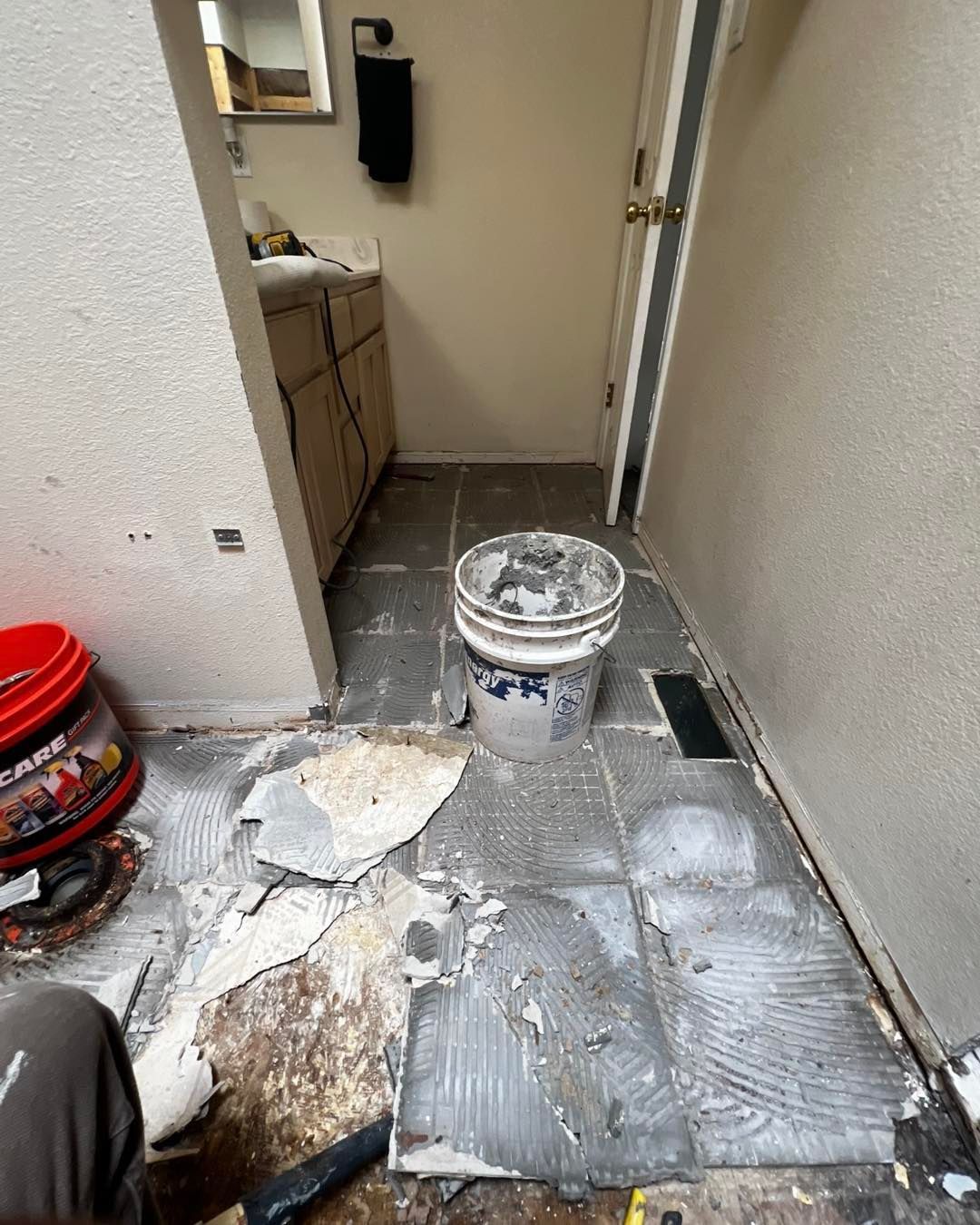 Bathroom floor with tile removal in progress, debris, a bucket, and a partially open door.