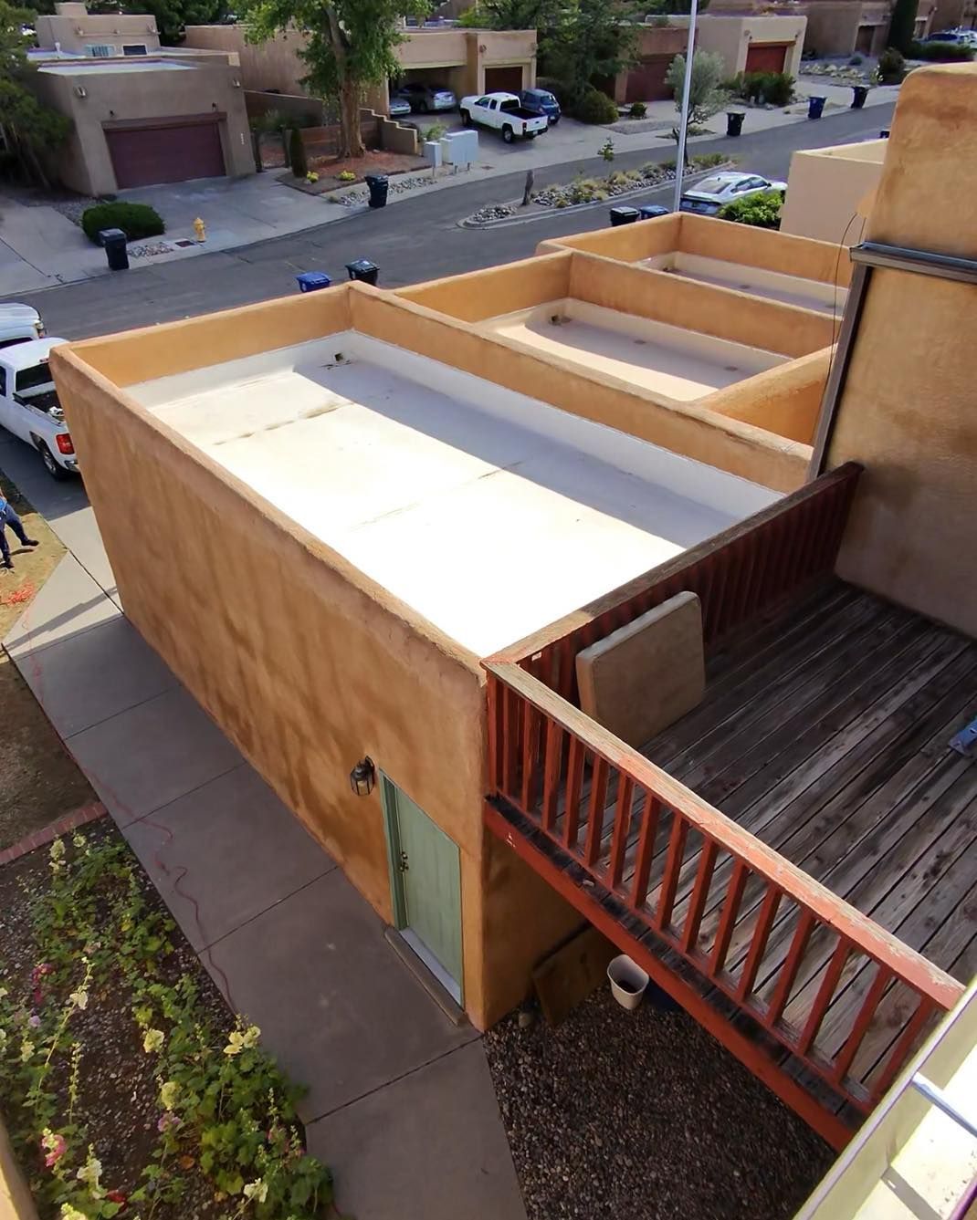 View of a flat-roofed stucco building with a wooden deck and walkway. A street and other homes are visible in the background.