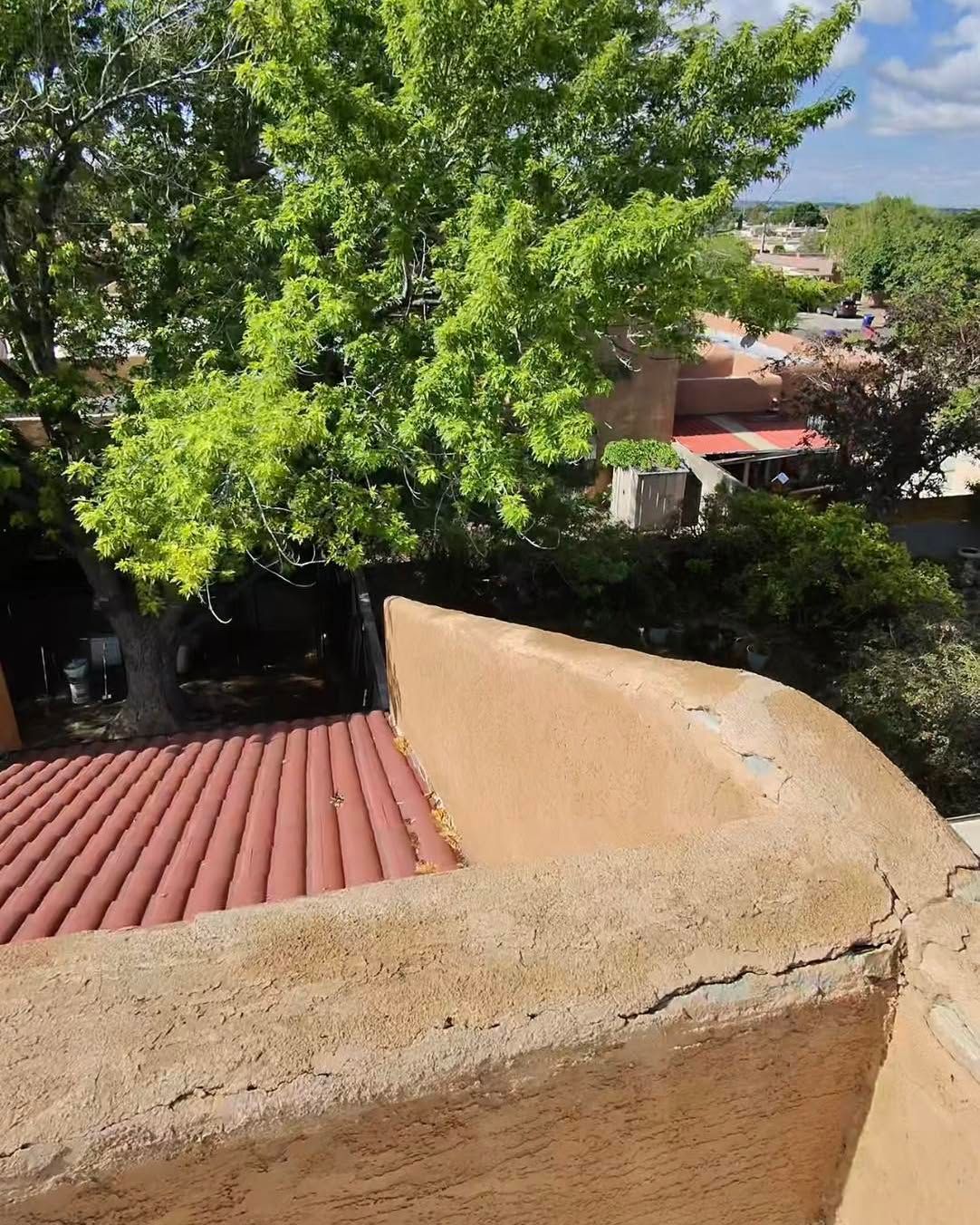 Tan stucco rooftop with red tile roof, tree, and distant buildings on a sunny day.