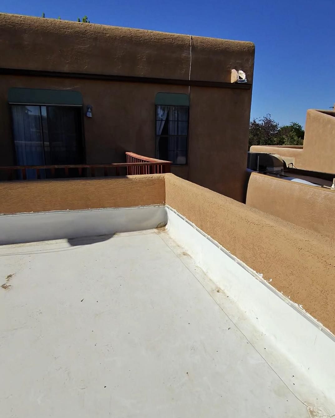 Exterior view of a stucco building with a flat roof. A rooftop balcony has a white painted edge and a short wooden railing.