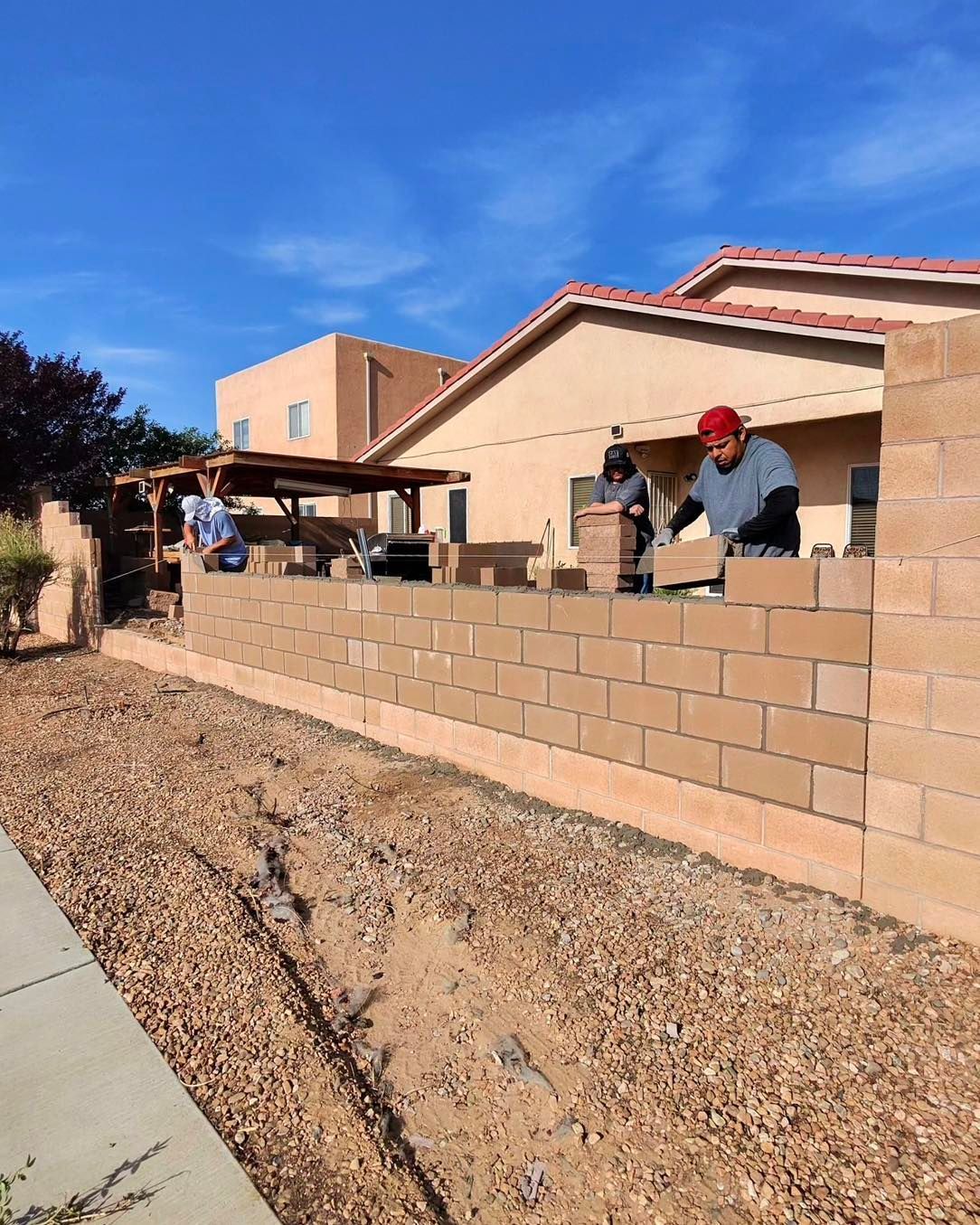 Construction workers building a cinder block wall outdoors under a blue sky, next to a home and a gravel area.