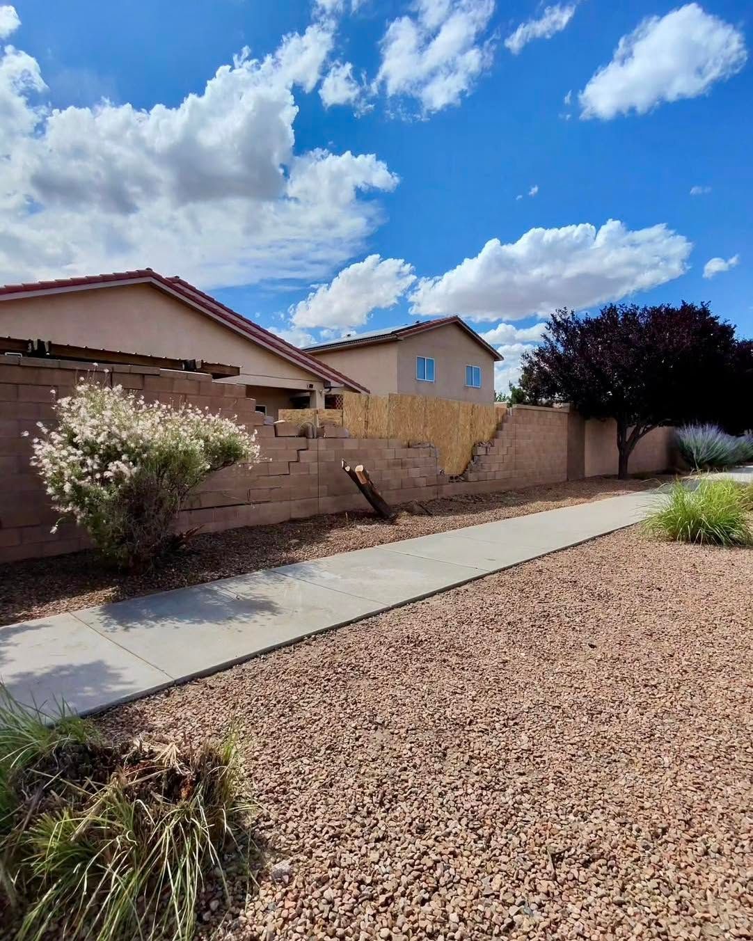 Sidewalk next to a brown gravel yard and wall, two houses, and a blue sky with clouds.