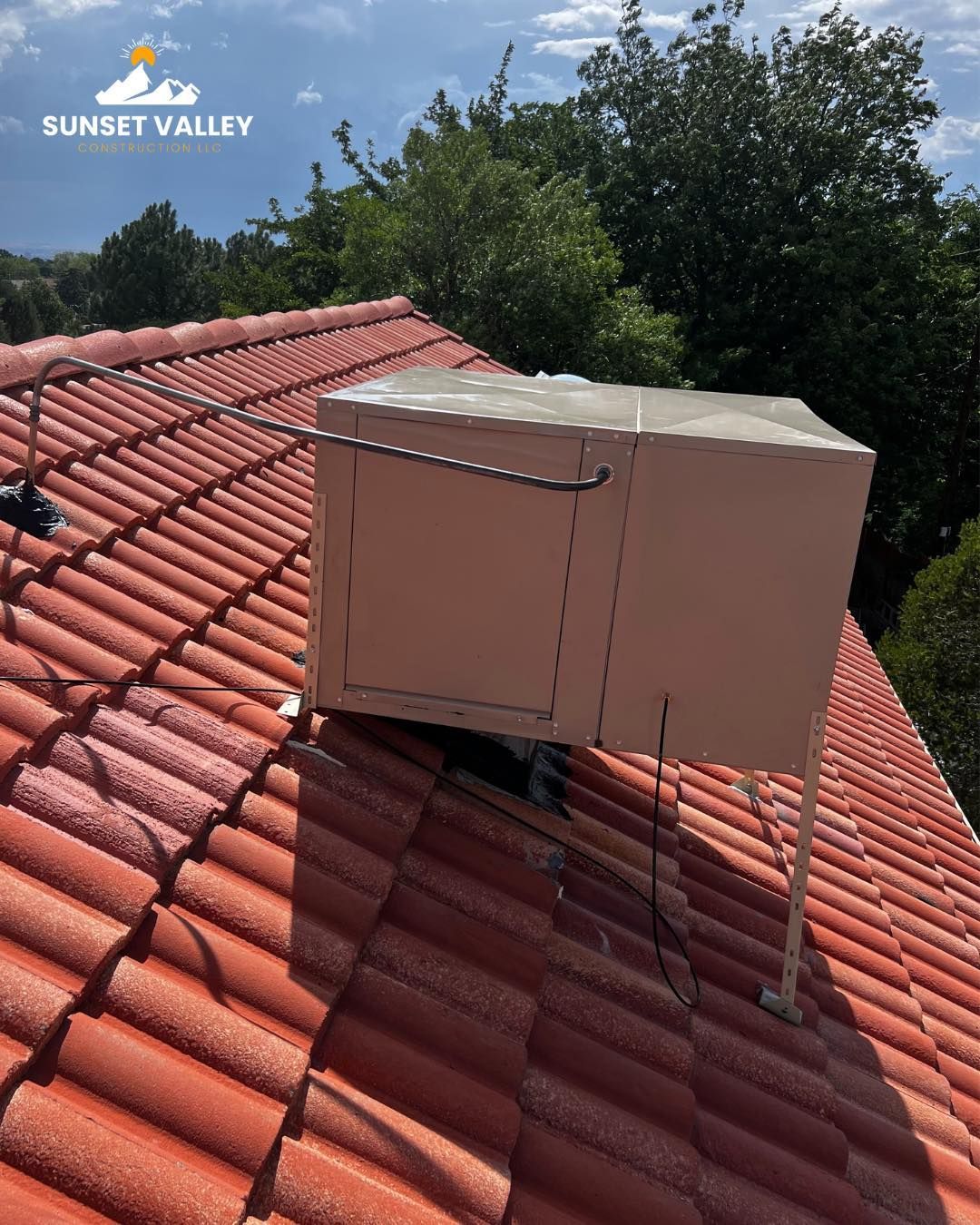 Large beige rooftop unit on a red tiled roof, with trees in the background under a blue sky.