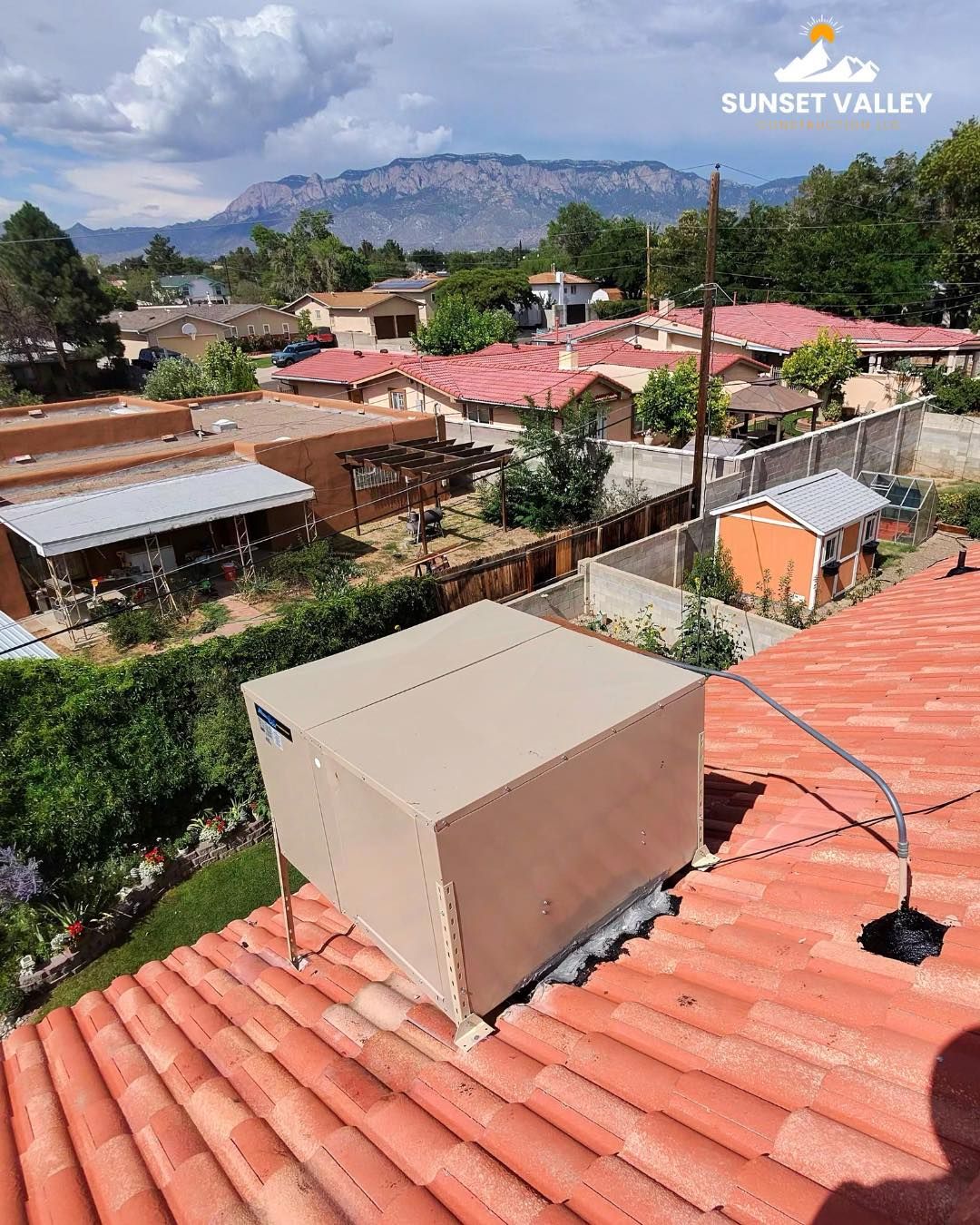 Beige box on a red tile roof, suburban view with houses, trees, and mountains under a partly cloudy sky.
