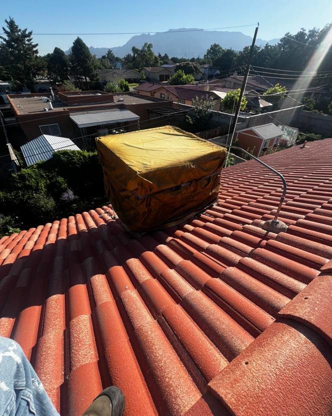 Red-tiled roof with a yellow, square object; a metal antenna and houses with a mountain in the background.