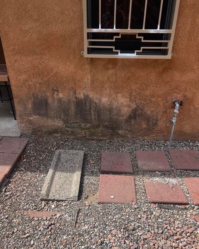 Exterior wall with water damage and a window with a metal grate, next to a water spigot. Ground covered in rocks and tiles.