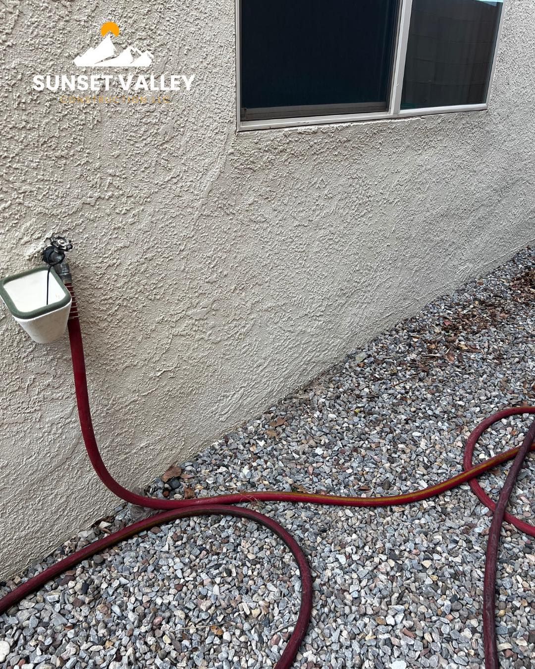 Exterior wall with spigot and red hose on gravel. A window is above.