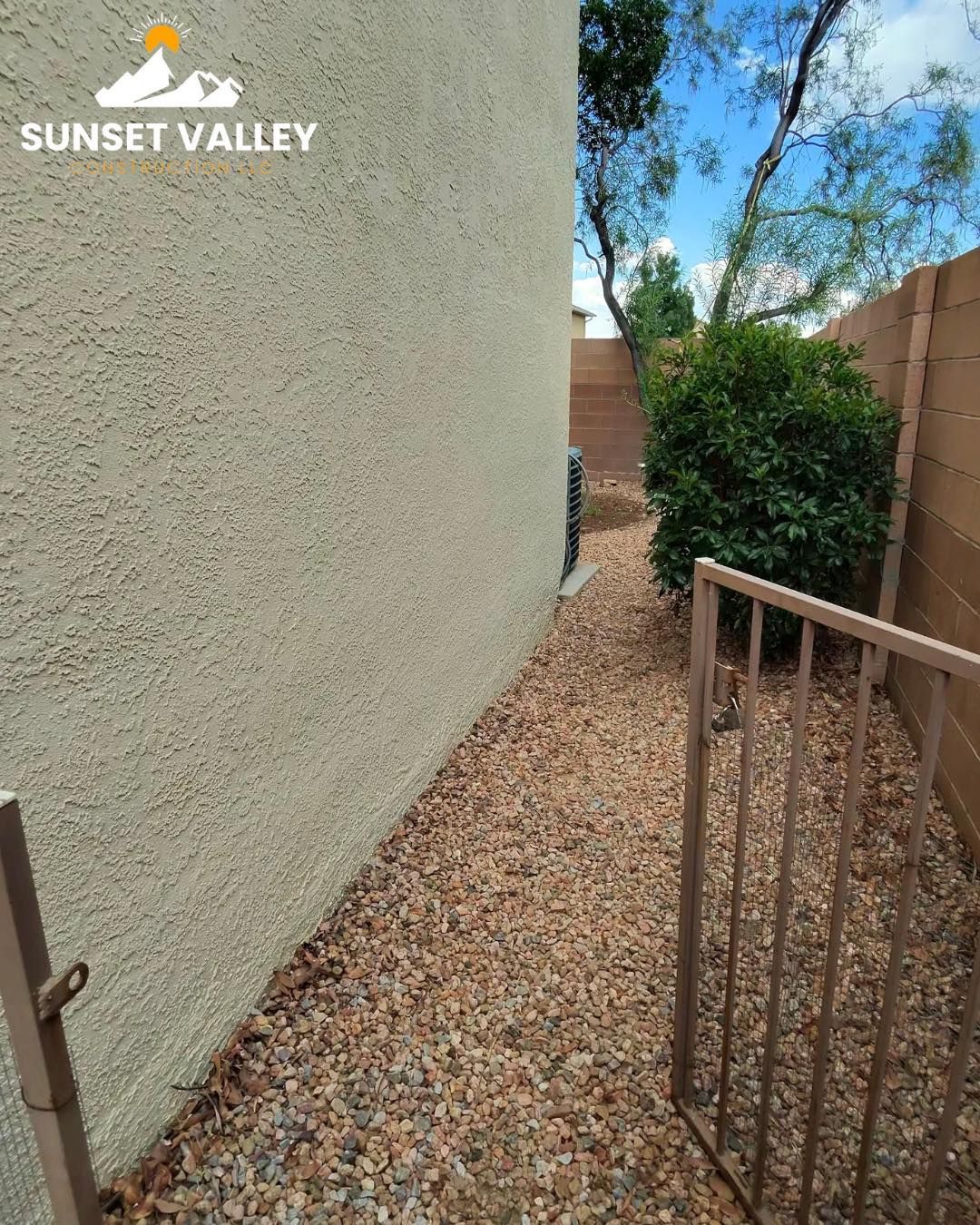 Narrow gravel path between a textured wall and a brown fence. Small gate open.
