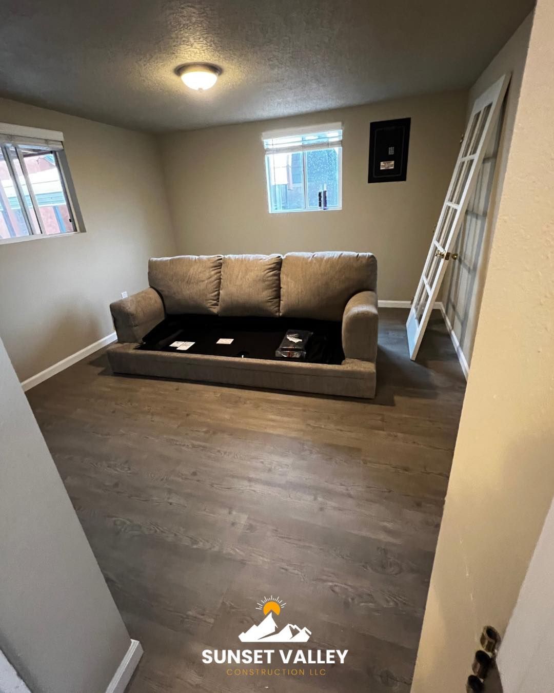 A room with a gray couch, two windows, and dark wood-look flooring. A door leans against the wall.
