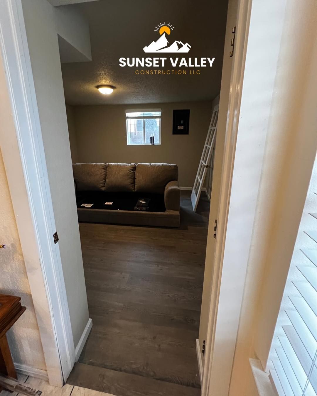 View into a room with a brown couch and a window. Wooden floor and a ladder visible.