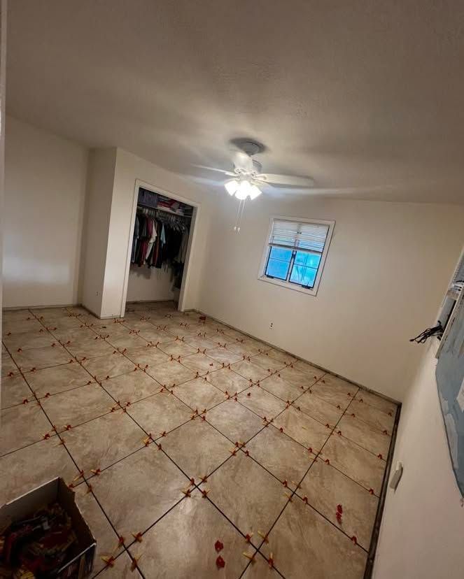 Bedroom with beige tiled floor, closet, window, and ceiling fan. Red tile spacers are visible.