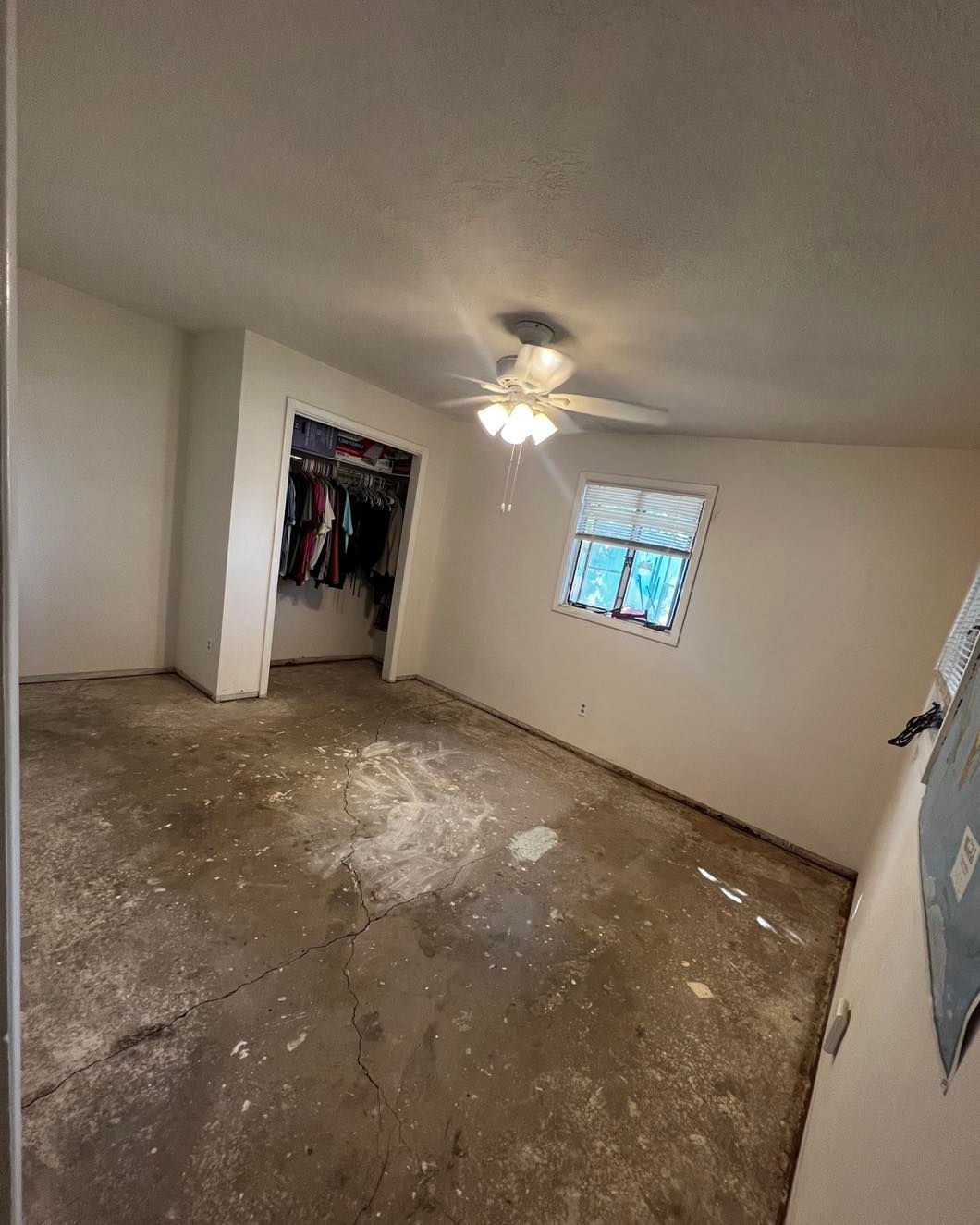 Empty room with damaged concrete floor, small window, open closet, and ceiling fan. White walls.