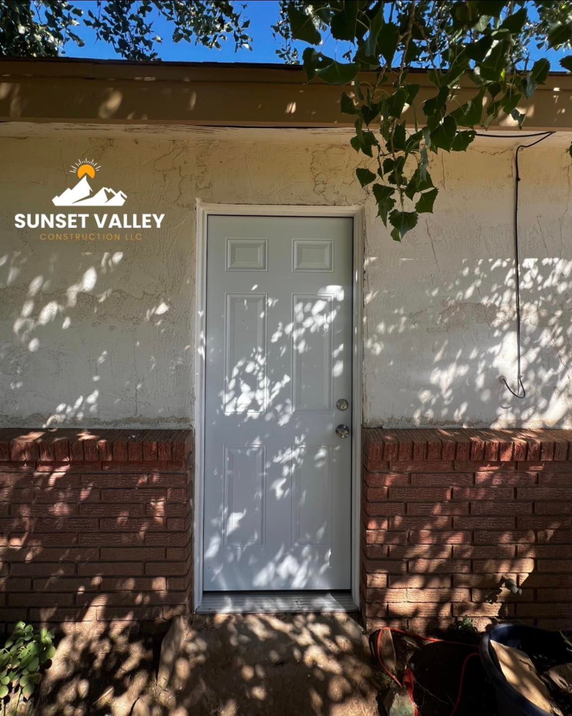White door on a stucco wall, shadowed by tree branches, with a brick base.
