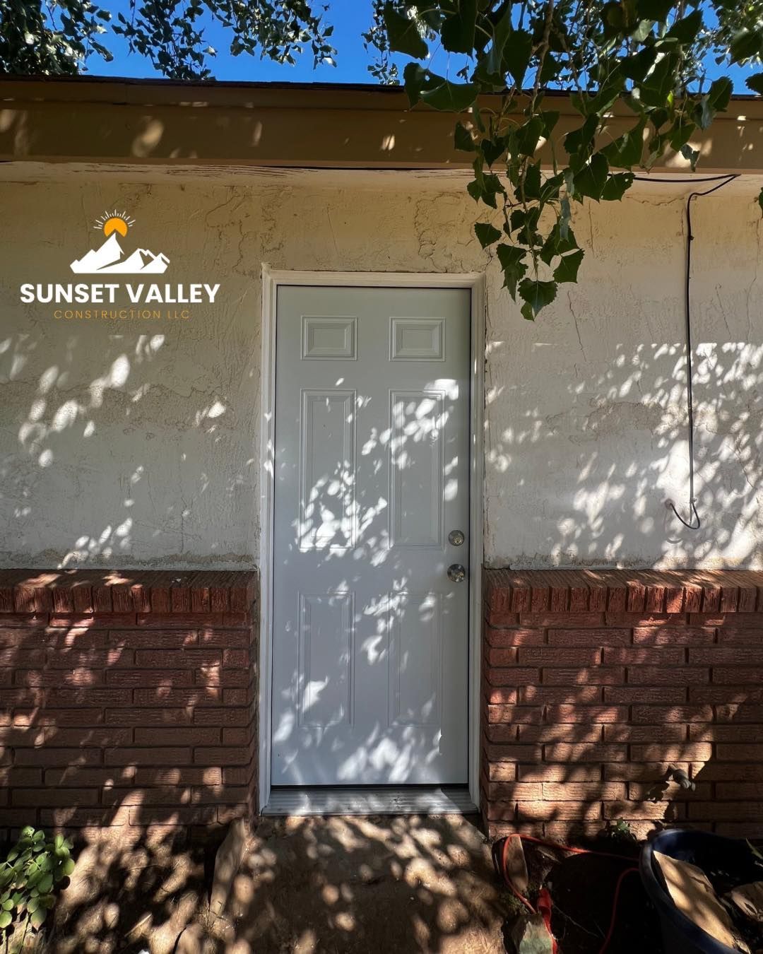 White door on a stucco and brick wall. Sunlight creates shadows from a tree. Sunset Valley logo.