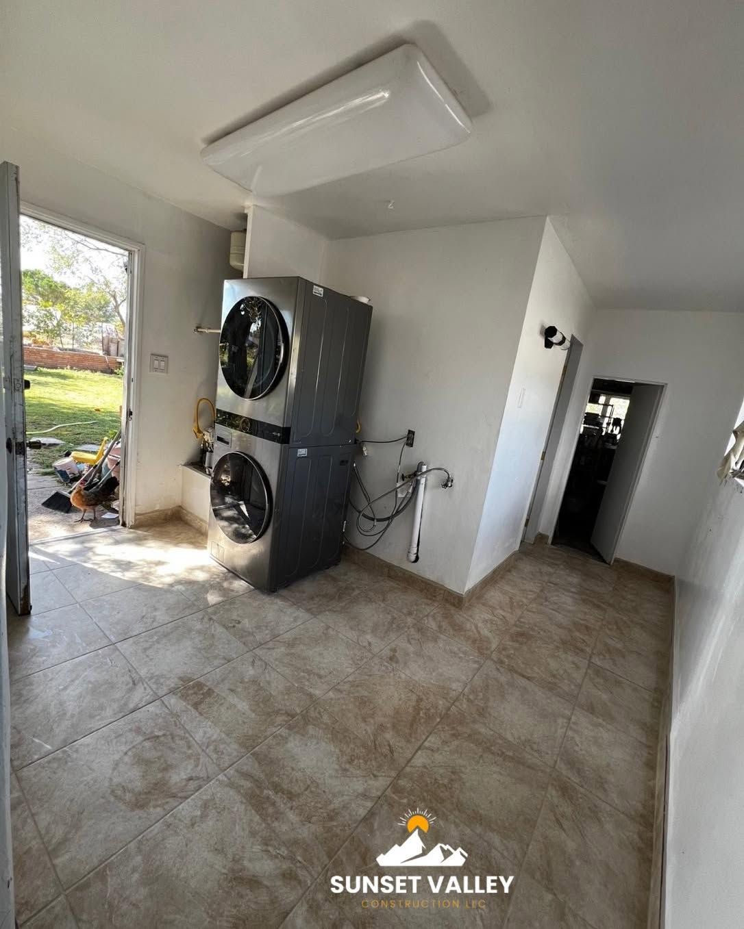 Laundry room with stacked washer and dryer, tile floor, open door to outdoors.