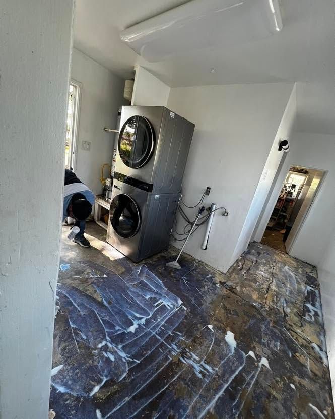 Stacked washer/dryer unit in a room with damaged flooring. A person is working nearby. Bright interior.
