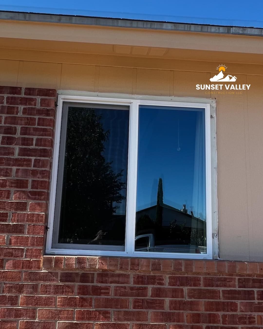 Window in a brick wall reflecting a building and a tree. White frame, beige siding, blue sky.
