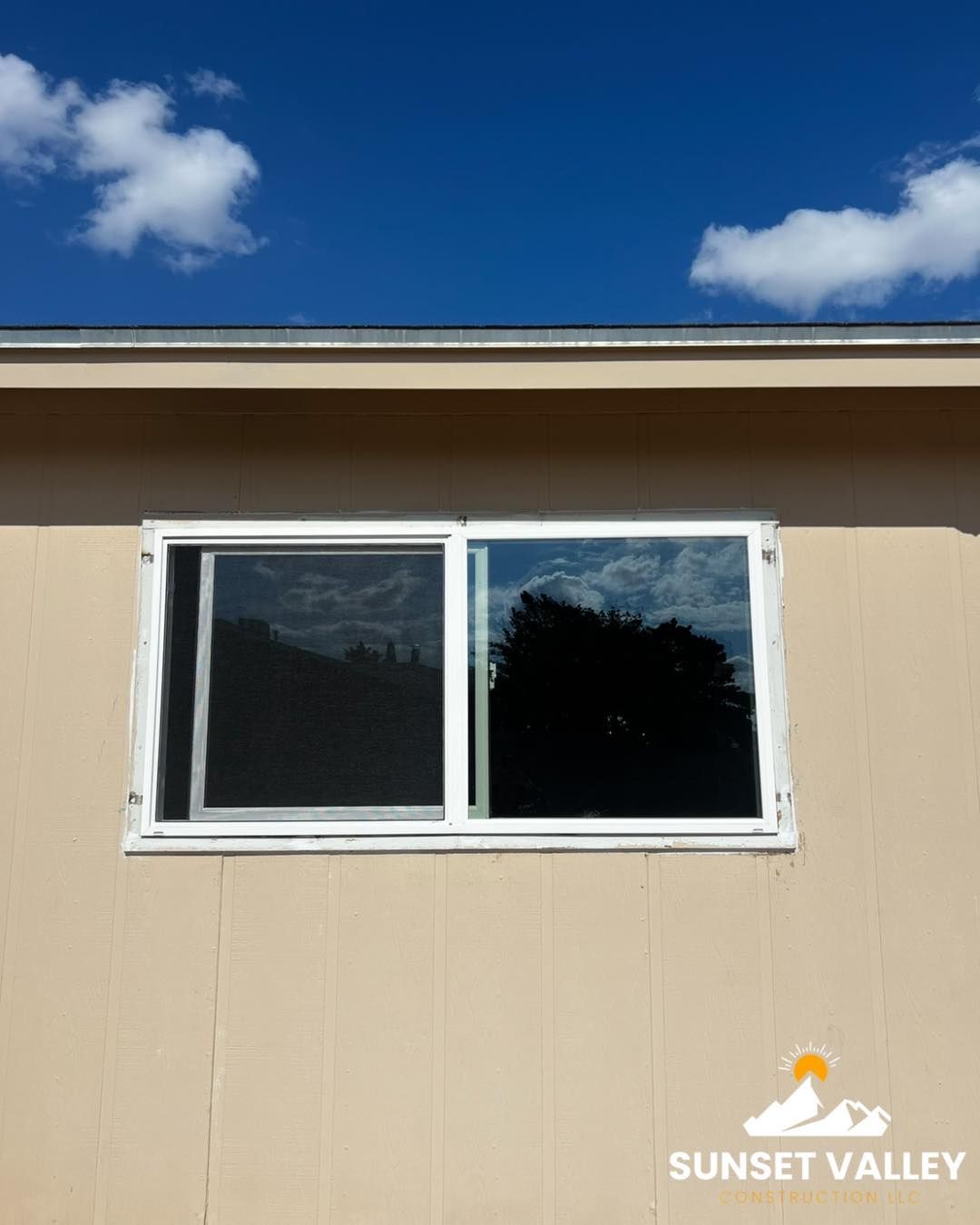 Window on beige building reflecting sky and tree, under a bright blue sky with clouds.