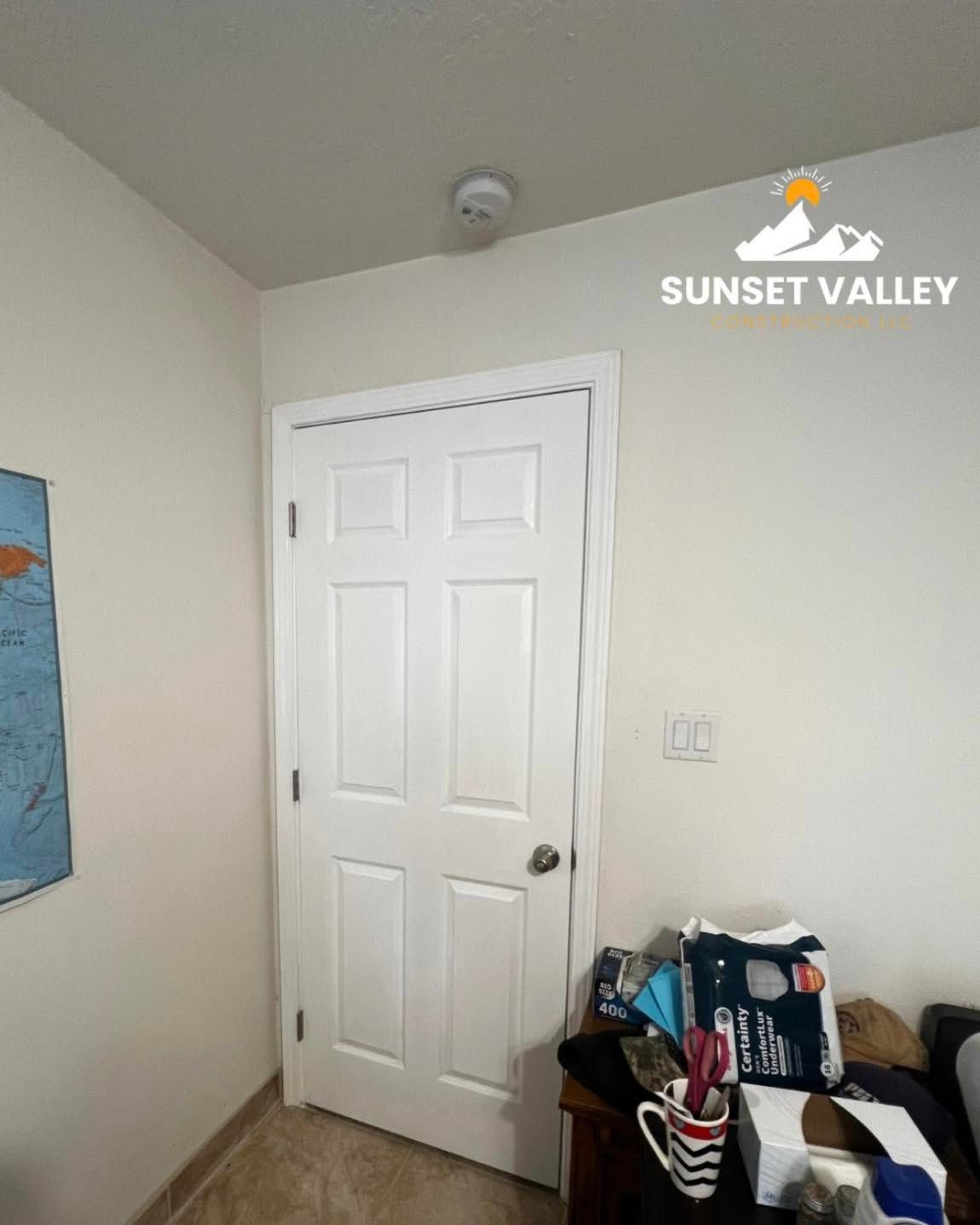 White door, smoke detector on ceiling, items on a nightstand, and a light switch on wall.