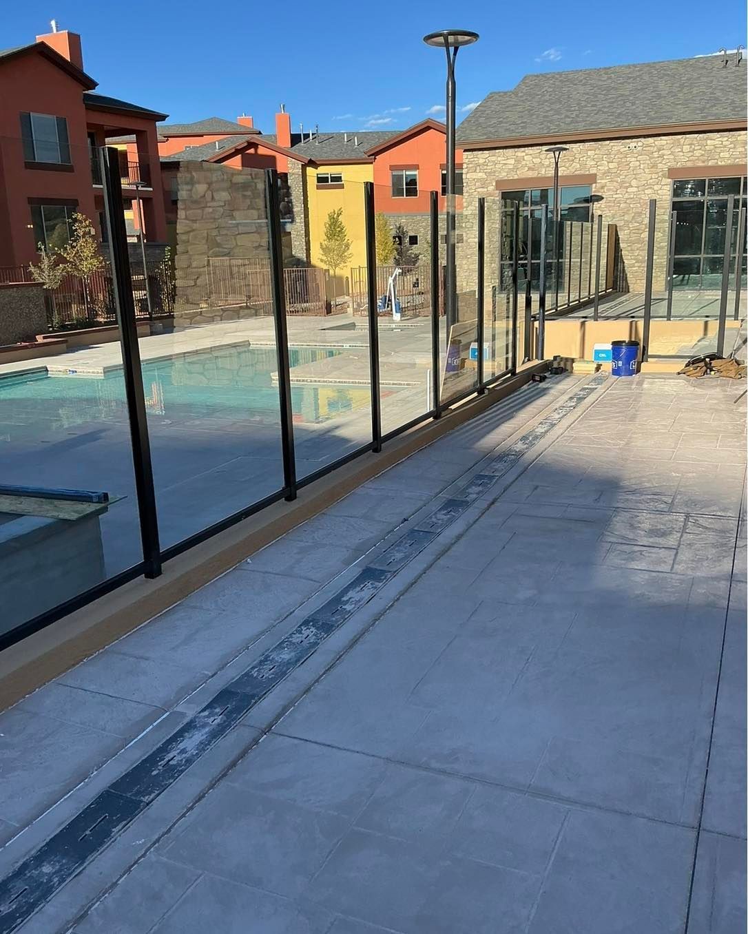 Pool area with glass fencing, concrete walkway, and colorful buildings in the background.
