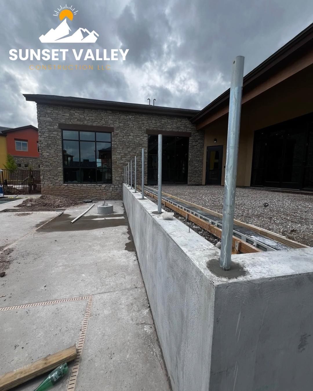 Concrete retaining wall with metal posts, under construction, near a building with stone facade.