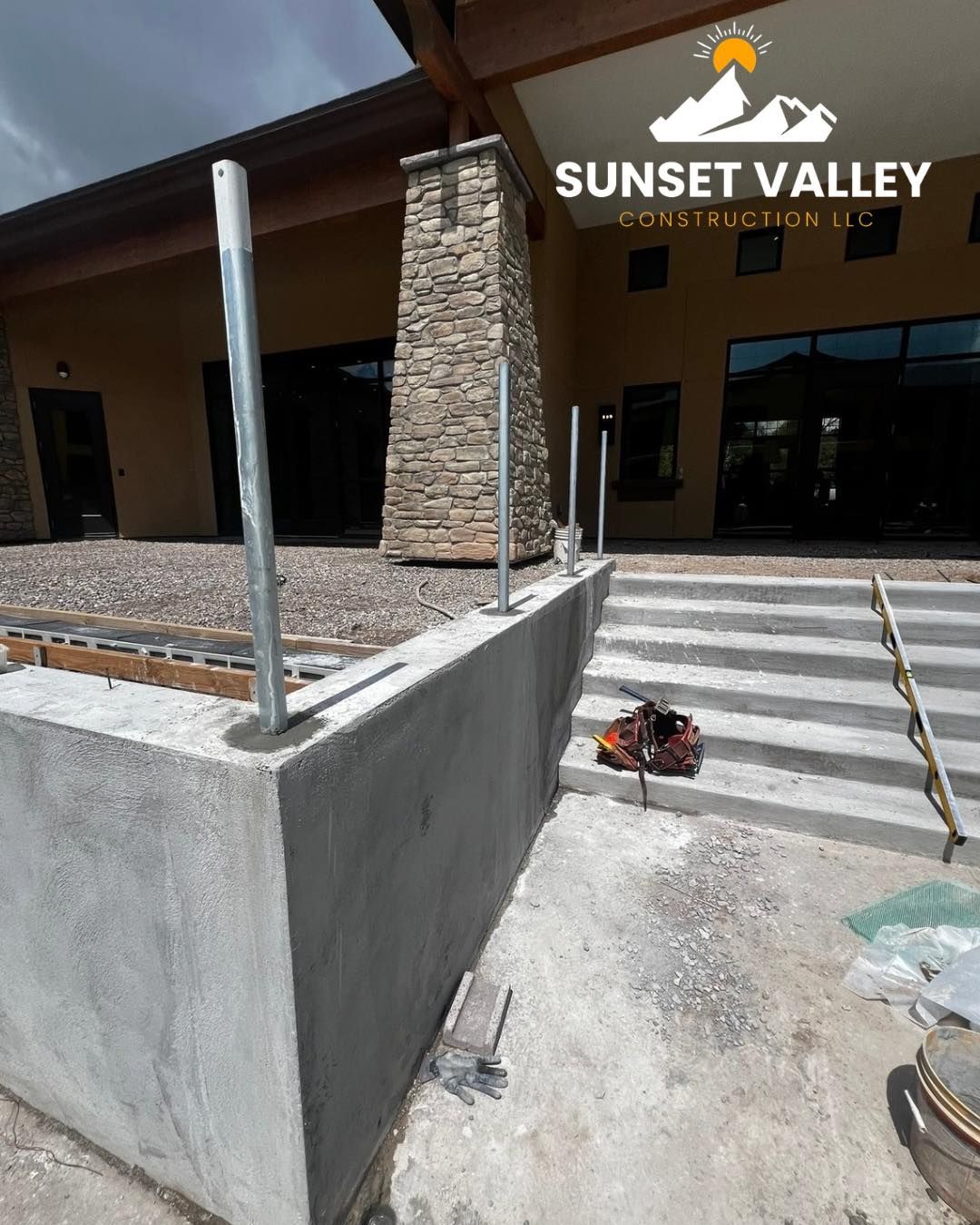 Construction site: concrete retaining wall with metal posts. Stairs lead up to a building with stone column.