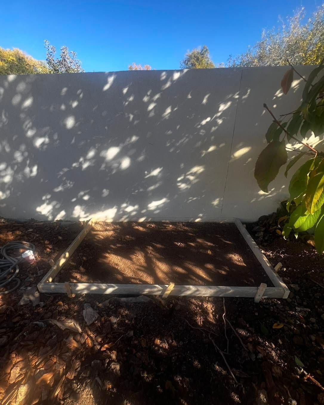 Concrete frame on dirt in front of a white wall, shaded by tree leaves, against a blue sky.