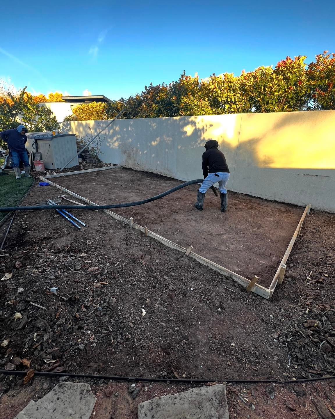 People working on a garden plot, bordered by wood, with tools and a hose, under a blue sky.