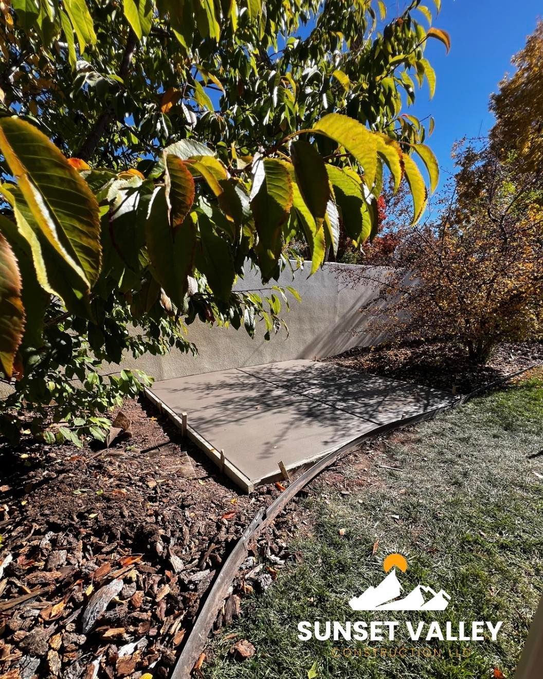 Concrete drainage channel in a grassy area, under a tree with yellowing leaves, against a blue sky.