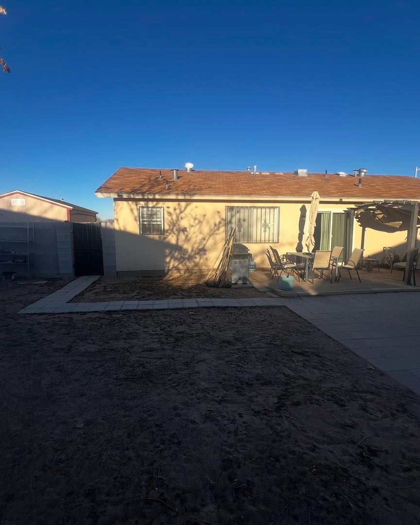 Backyard with a tan house, brown roof, and blue sky. A concrete path and a shed are also visible.