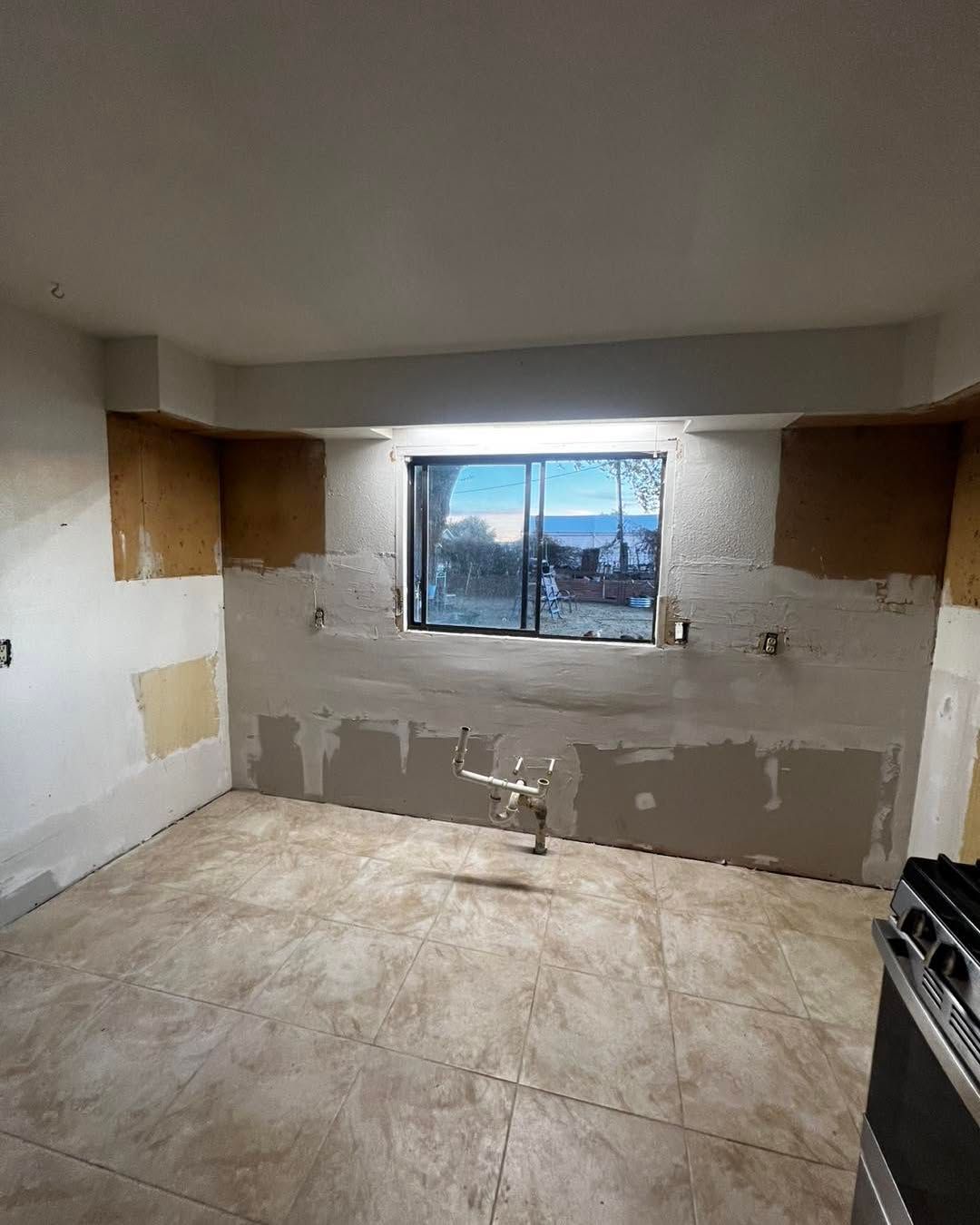 Kitchen under renovation; walls with exposed drywall, tile floor, window with a view, and gas stove.