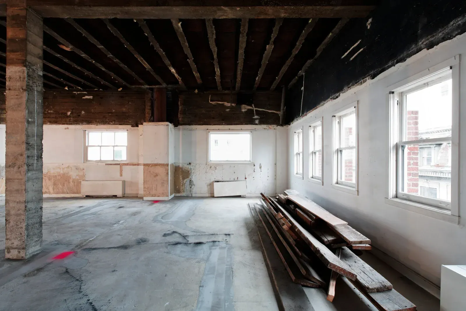 Interior of a building under renovation with exposed beams, concrete floor, and windows; lumber stacked on the floor.