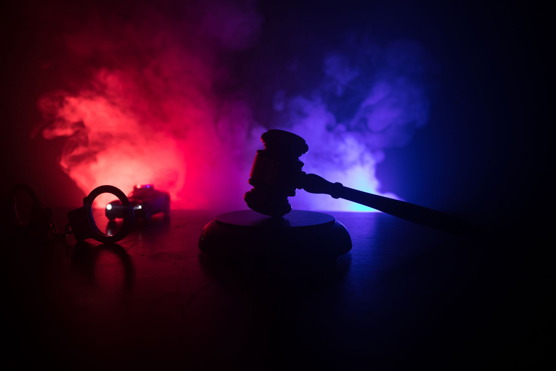 Gavel on block with handcuffs silhouetted against red and blue smoky background, representing law and justice.