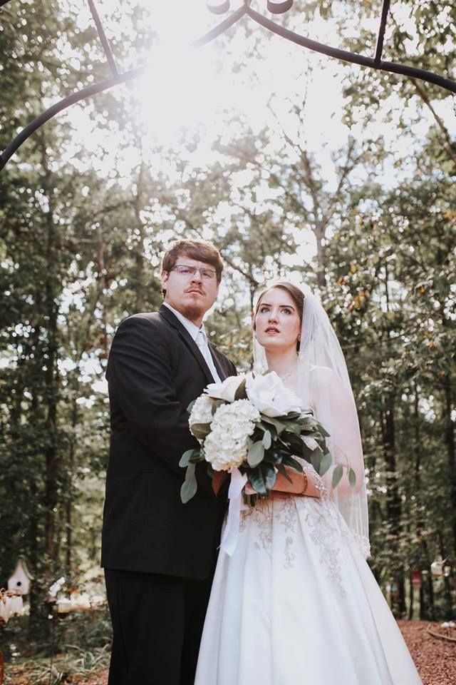 Bride and groom in formalwear holding a bouquet in a sunlit forest ceremony