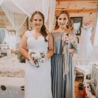 Two women in formal dresses standing together indoors, one in white holding a bouquet and one in gray holding flowers