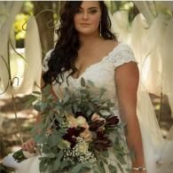 Bride in white lace dress holding a greenery bouquet outdoors under a floral arch